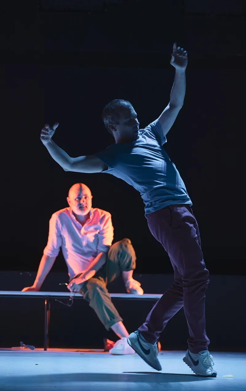 Dans « Scroll », un comédien en plein mouvement devant un homme assis sur un banc, sur un plateau sombre.