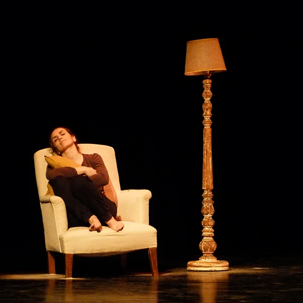 Dans « Madame Magarotto », une comédienne recroquevillée dans un fauteuil clair, près d’un lampadaire doré, sur fond noir.